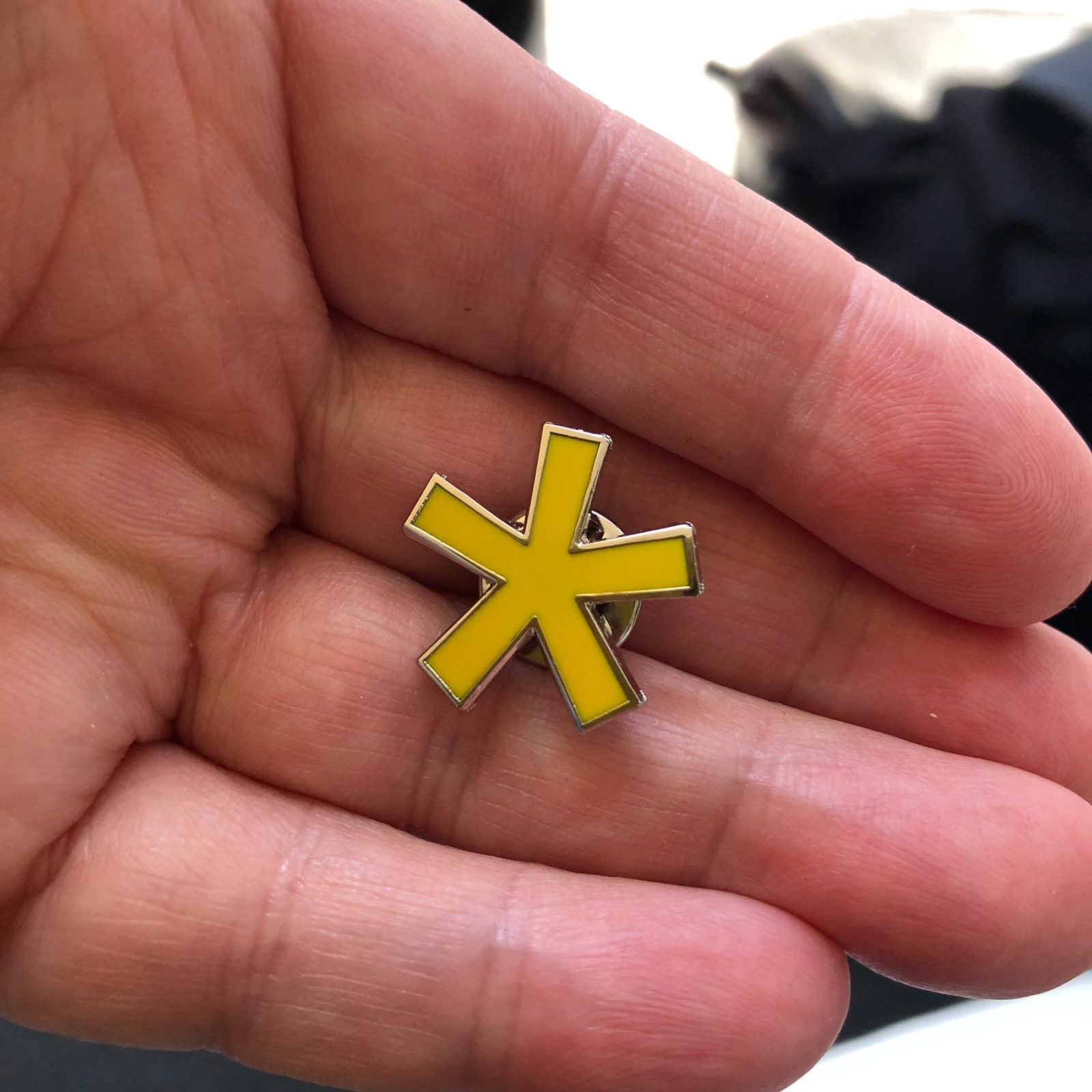 A Signal Yellow asterisk enamel pin held in a hand